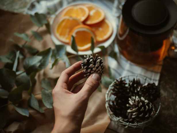 hand holding pinecone above tea pot and orange slices on plate